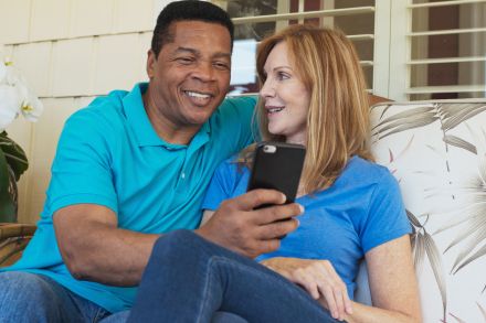 Positive diverse couple in casual clothes sitting at table with cup of drink and eating delicious food while spending time together in kitchen at home